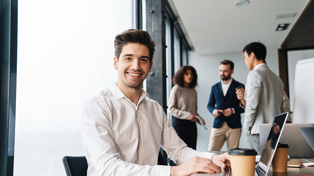 Attractive young confident businessman sitting at the office table with group of colleagues in the background, working on laptop computer